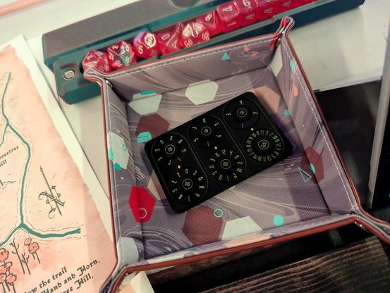 A thick black metal card with 6 individual dials, each representing a D4, D6, D8, D10, D12, and D20, as represented in tiny yellow text. The card sits in a small dice tray on a white table next to a hand-drawn map and a dice box holding a full set of glittery pink fantasy dice.