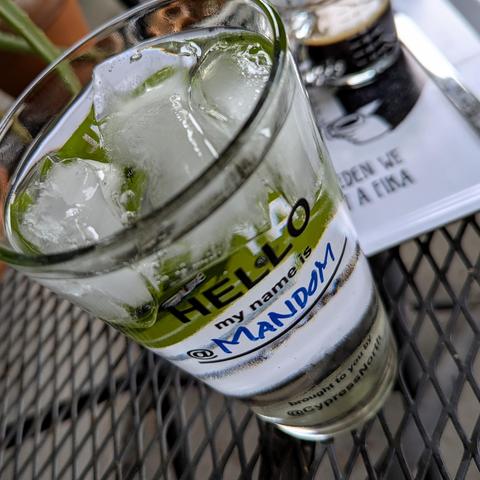 A pint glass printed with “Hello my name is,” and hand-written in marker below it is, “MANDOM.” In the background is a white tray,a shot of espresso waiting to go into the club soda and ice in the pint glass, and a bowl of pretzels.