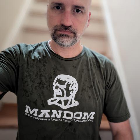 Selfie of a shaved bald white guy with a graying beard wearing an olive tee shirt showing a brutalist line-art representation of Charles Bronson’s blocky head with shaggy hair and thin mustache. Below that in bold letters is ”MANDOM” and below that is smaller text, “All the world loves a lover. All the world loves MANDOM.”