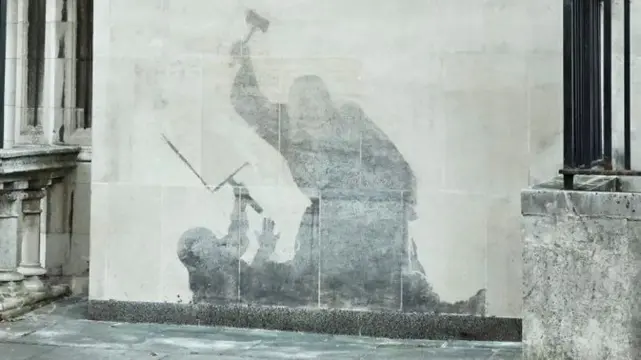 A stone court building exterior wall showing the silhouette of a person on the ground holding up their hand and a protest sign in a defensive posture as a robed figure looms over it, gavel on the backswing to strike. The silhouette is inverted, in that the rest of the stone is caked in lighter grime, but the silhouette shows freshly cleaned pristine stone.
