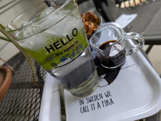 A pint glass filled with ice, fizzy, and simple syrup, alongside a shot glass of espresso and bowl of pretzels, all sitting on my fika tray; written on the glass in marker is #id24.