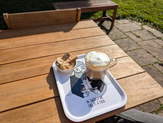 A foamy cappuccino in a glass mug next to a tiny bowl with equally tiny biscotti, on my fika tray, on a small wooden table, on a stone patio in a grassy yard, with a low sun making stark long shadows.