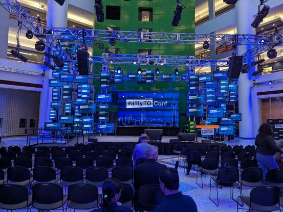 A 10 atory atrium with a stage backed up to a green wall of elevators, surrounded with trusses and framing holding around 60 televisions of assorted sizes and orientations, loosely collected around a main presentation screen.