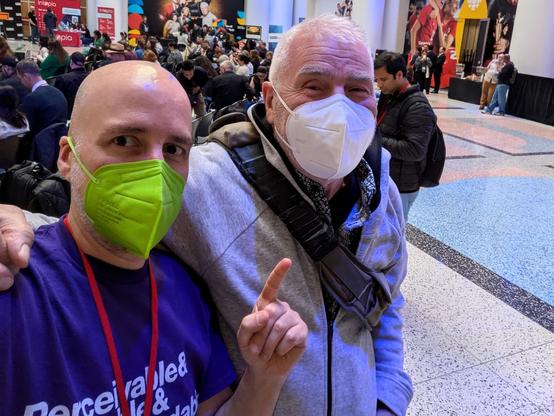 Two old white guys, one with a shaved head and beard in a green mask, the other with short white hair and beard in a white mask, posing for a photo as I snapped a selfie.