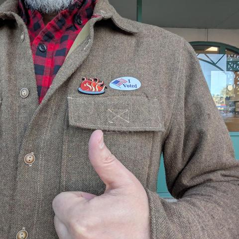 Selfie showing just my chest with an “I voted” sticker next to a pin with a cooked tentacle ham on a brown herringbone jacket as I give a thumbs-up.