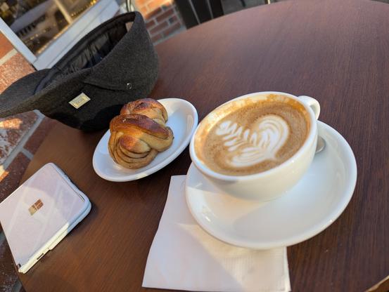 Outdoor table with a low sun lighting a cappuccino in a porcelain mug along with a knotted dough treat, both next to a Surface Duo and a fancy brimmed wool hat and brick wall.
