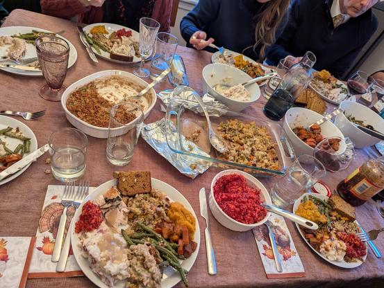 Tight shot of the table with a bright red cranberry relish, a glass dish of colorful stuffing, a white ramekin with crispy green bean casserole, a metal tray of brown bread showing with pecans and cranberries in the cross section, white bowls holding mashed potatoes, green beans with crispy crumbs, and dark orange squash and carrot chunks; the plates are filled and a jar of chili crisp sits off to the side.
