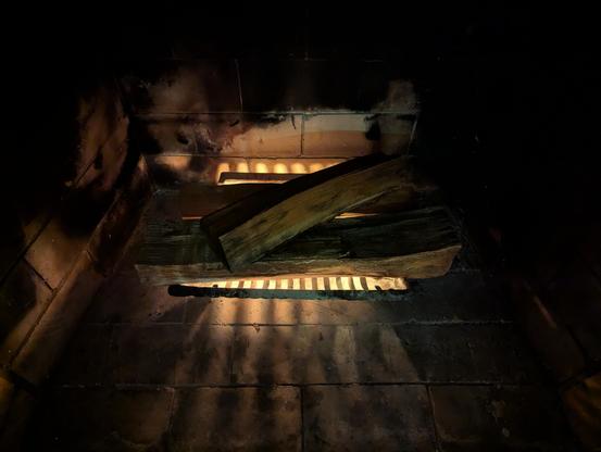 Looking down over four logs in a dark fireplace, orange light coming from below the logs as bright fingers on the ground thanks to shadows cast by the iron grate as the fire starter sticks start to burn.