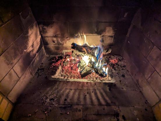 Looking down again, but the logs are reduced to bright red coals and a few large chunks of flaming charcoal, with spillover of embers and ash around the grate and loose crumbs of black further out.