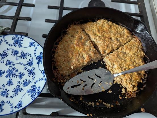 A 10 inch cast iron skillet with four wedges of baked rolled oats, one missing, showing a very rough texture and golden browning on top.