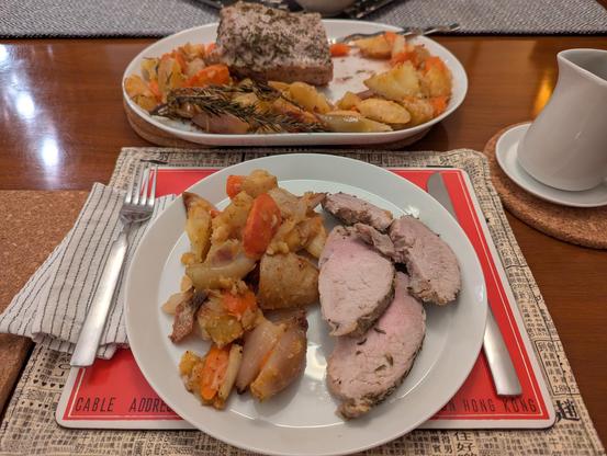 A large plate with an assortment of roasted vegetables and very pink slices of pork loin, with a platter of pork roast and vegetables in the background and a small pitcher for gravy.