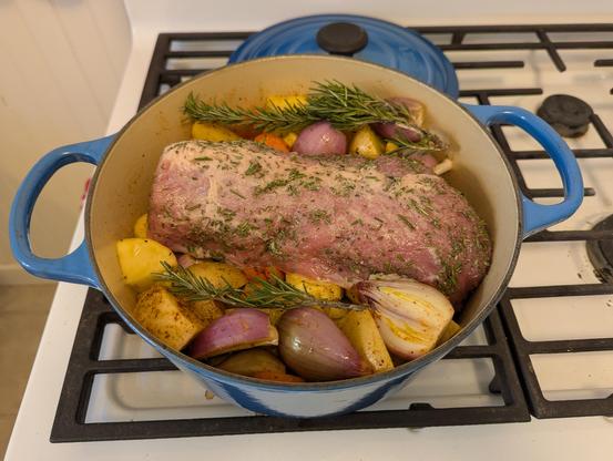 A Le Creuset Dutch oven filled with potatoes, carrots, parsnips, shallots, rosemary sprigs, and a large bonelss pork loin encrusted in salt and chopped rosemary, all before going into the oven.