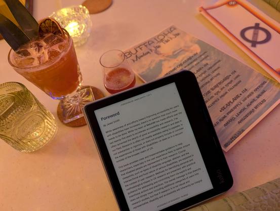 A kobo e-reader on a white bar with a drink menu labeled “Buffaloha Monday Nite Tiki Nite” from Nowhere Lounge, and behind the e-reader is a small 4 ounce glass of something dark pink, a bigger chunky and textured glass with two pineapple leaves sticking out of ice and another dark pink drink, and a greenish textured tumbler of water.