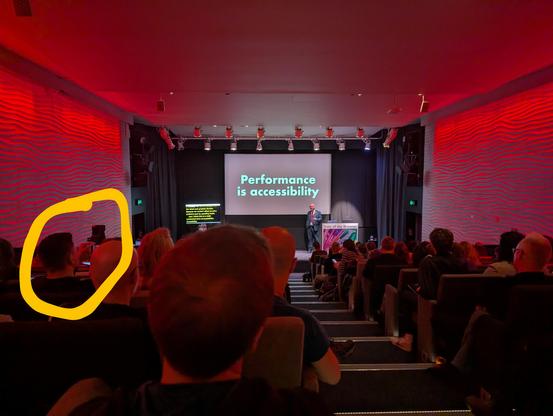 “Performance is accessibility” projected on a stage in a long and narrow auditorium full of people, one of whom is circled in yellow, with his phone raised to take a photo of the slide.