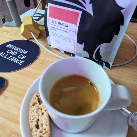 Looking down into an espresso cup half full of espresso, with a tiny biscotti and spoon on its saucer; a bag of whole bean coffee labeled La Bermeja (forest fruits, plum, almond) with a stylized gryphon silhouette.
