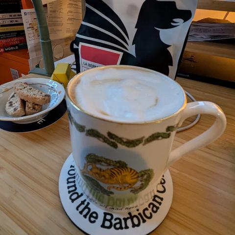 A fine china mug painted with a Cheshire cat and the text “We’re all mad here,” filled to the top with milk foam, sitting on a Barbican coaster; a few biscotti and the bag of beans visible in the background.