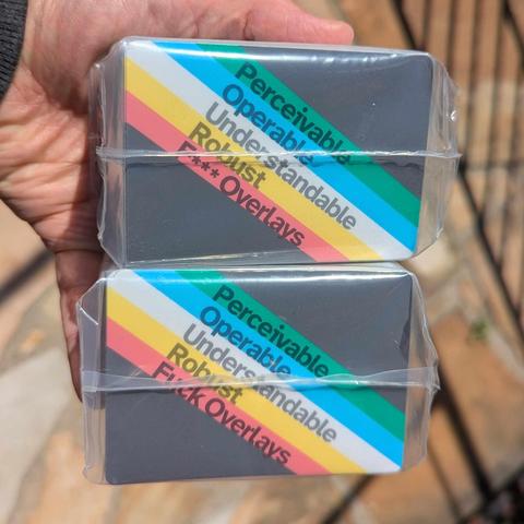 Holding two packs of 3½ × 2½ inch stickers — a dark gray field with five light stripes going from the upper-left to bottom-right corner: green, blue, gray, yellow, and red; within each stripe is a word: Perceivable, Operable, Understandable, Robust, F*** Overlays. The second set of stickers has the word ‘Fuck’ spelled out.
