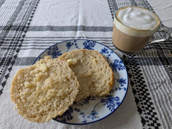 One of the baked rolls, sliced horizontally in half and buttered, sitting on a white plate with blue flowers next to a glass mug of cappuccino (with a nice, thick, white foam).