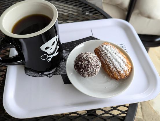 A golden madeleine dusted with powdered sugar alongside a large dark ball dusted in coconut, both on a saucer next to a black diner mug of coffee with a Jolly Roger in white but made from a cappuccino, a portafilter, and a wrench.