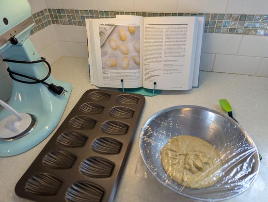 A metal bowl with yellow batter covered in plastic wrap next to a madeleine pan and in front of a cookbook with the recipe.
