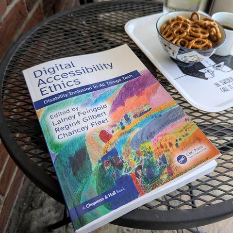 A softcover copy of ‘Digital Accessibility Ethics’ on a black metal mesh outdoor table alongside a small bowl of pretzels, an espresso cup, and a pint glass of iced club soda, all on my fika tray; the book is 7 by 10 by ¾ inches.