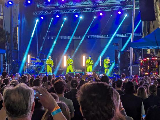 Outdoor stage in the evening; four men in a line across the front and one on drums, all wearing bright yellow janitor or hazmat suits, blue light beams sweeping over them.