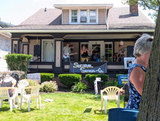 A deep shaded bungalow front porch with a guitarist, drummer, bassist, and singer.