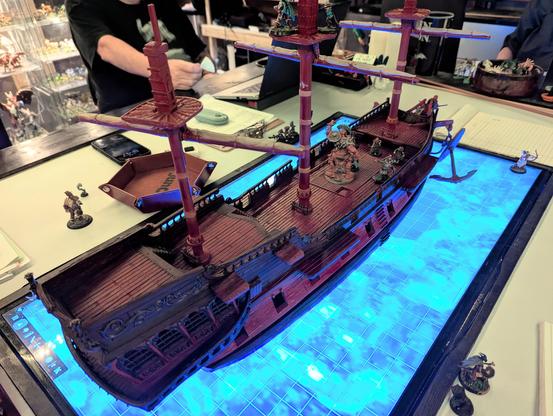 A model of a three-masted galleon in scale for 28mm miniatures, painted with some detail work, sitting on television screen embedded in a table, showing blue ocean water.