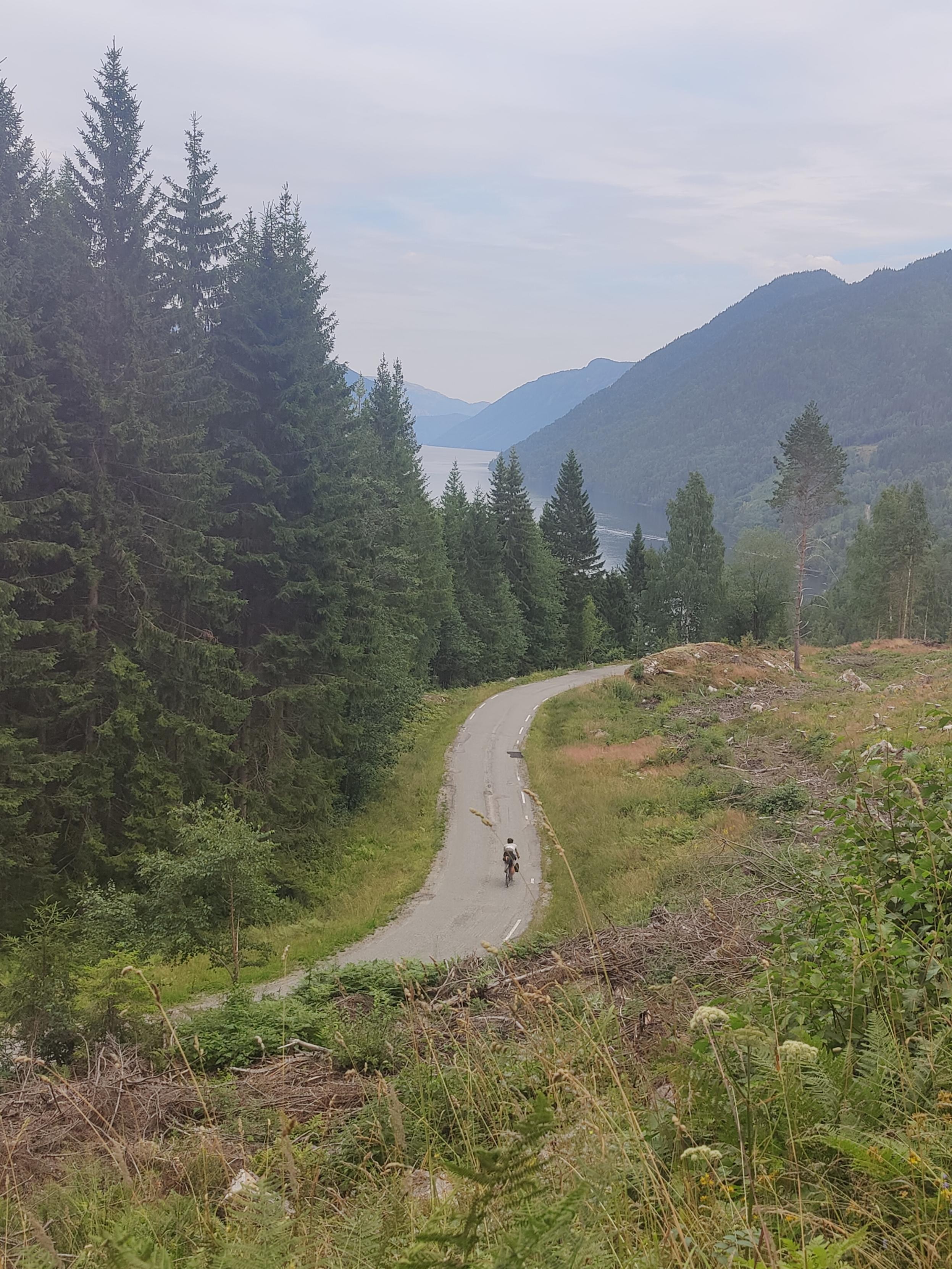 Cyclist descending on asphalt road, lake between mountains visible in the background.