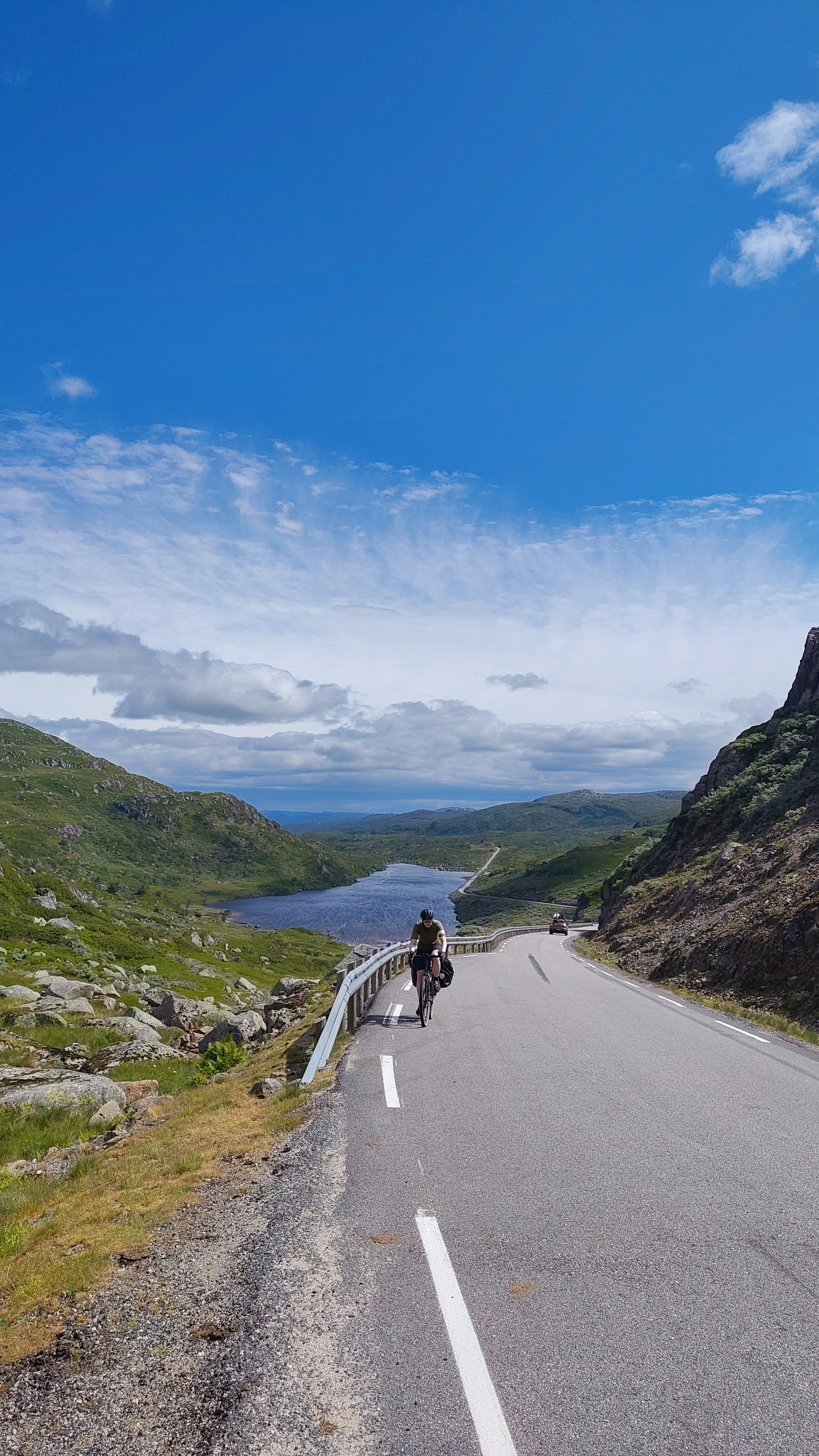 Cyclist on a mountain road, lake visible in the background.