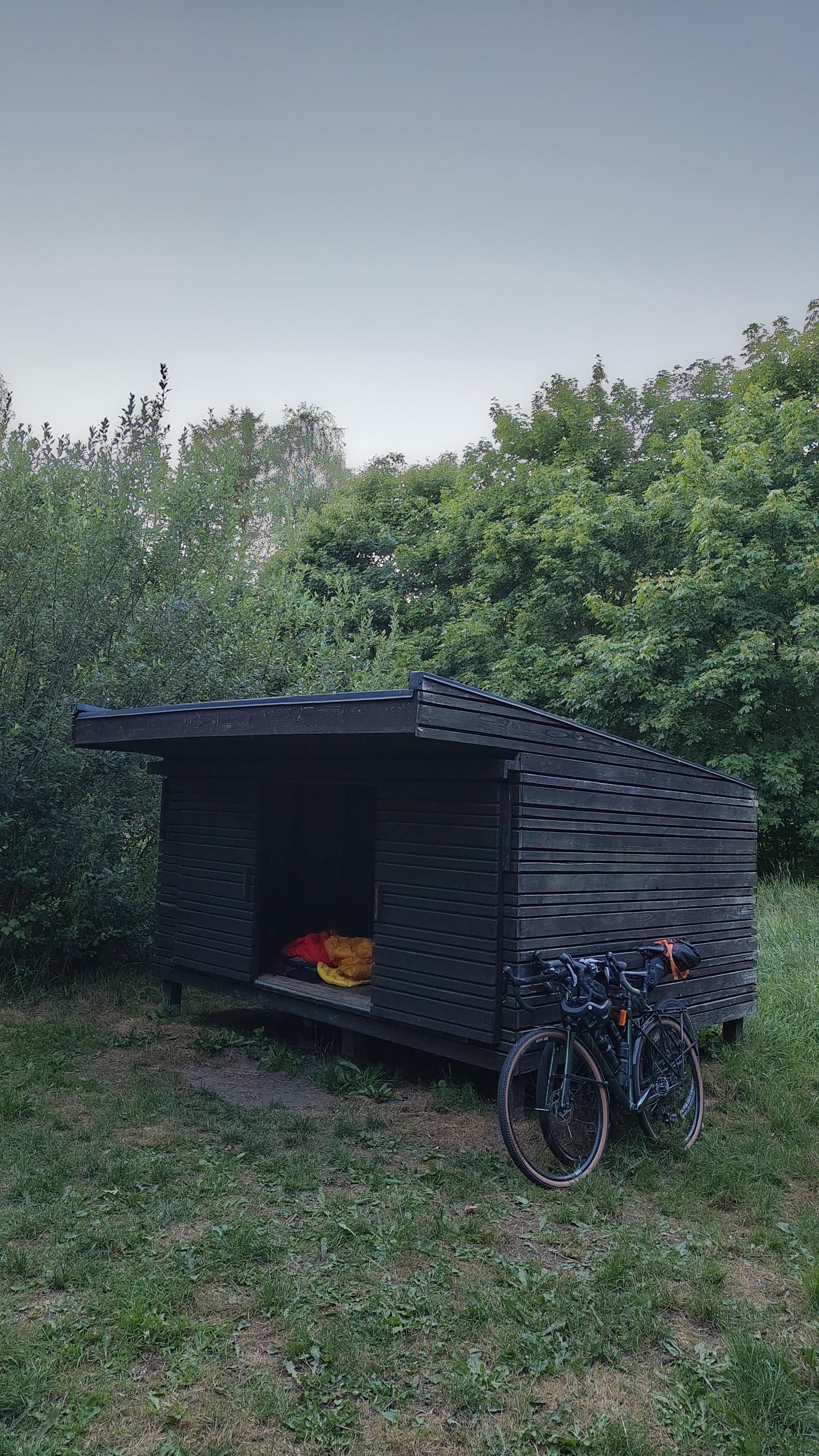 Two bikes next to a Danish shelter, which has the doors opened and some camping gear inside.