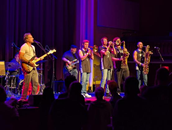Stage with a guitarist, drummer, bassist, trombonist, trumpeter, tenor saxophonist, and baritone saxophonist mid-performance; the horn section are wearing numbered jerseys with ‘Part Time Genius’ on them.