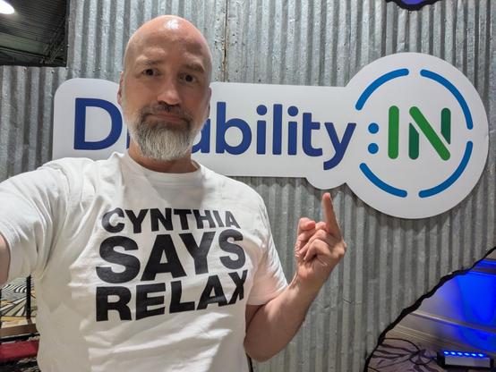 Selfie of me in front of corrugated metal Texas-shaped sign with Disability:IN logo, and I’m wearing a white tee with black all-caps text reading, “CYNTHIA SAYS RELAX”. I’m looking into the camera but holding up my middle finger off to the side.