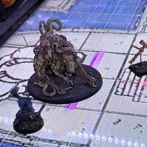 A painted gaming mini of a large (15 feet in game terms) fleshy creature with a mostly intestine-like body, tentacles, grasping elongated arms, and no discernable head, resting on a gaming map on a horizontal television screen.