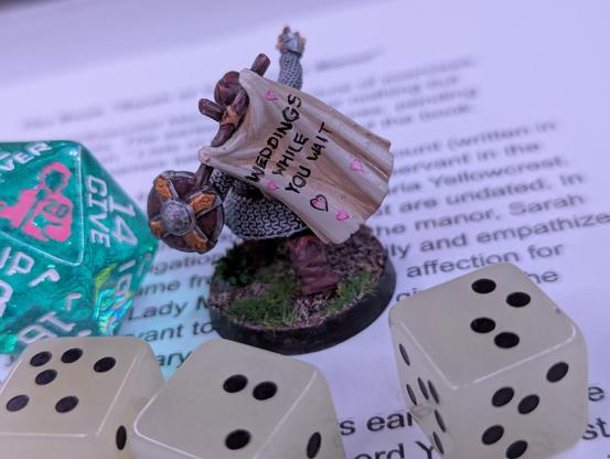 The same cleric mini, but now the tabbard on his back is visible and it reads, “Weddings while you wait,” which is surrounded by hearts.