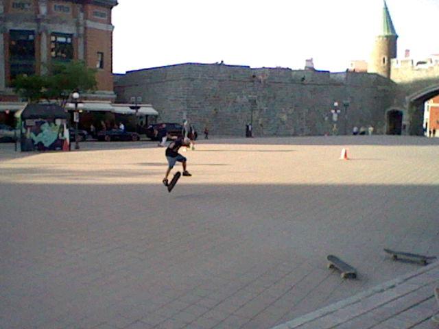 A large paved walled courtyard where a skateboarder preforms a kickflip in the late afternoon.