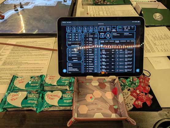 Four Treats, each with a hand-written note in a designated area of white: combat treat, crit treat, encounter treat, save treat. The are next to an iPad mini (showing a character sheet) on a stand over a small dice tray, all on a bigger table.