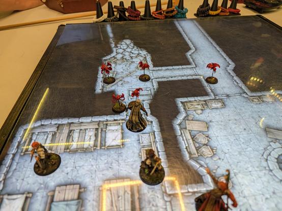 A table with a television set into it showing a map of tunnels and rubble, with a few character minis being swarmed by small, red, four-winged, spiked-nose baddies 
